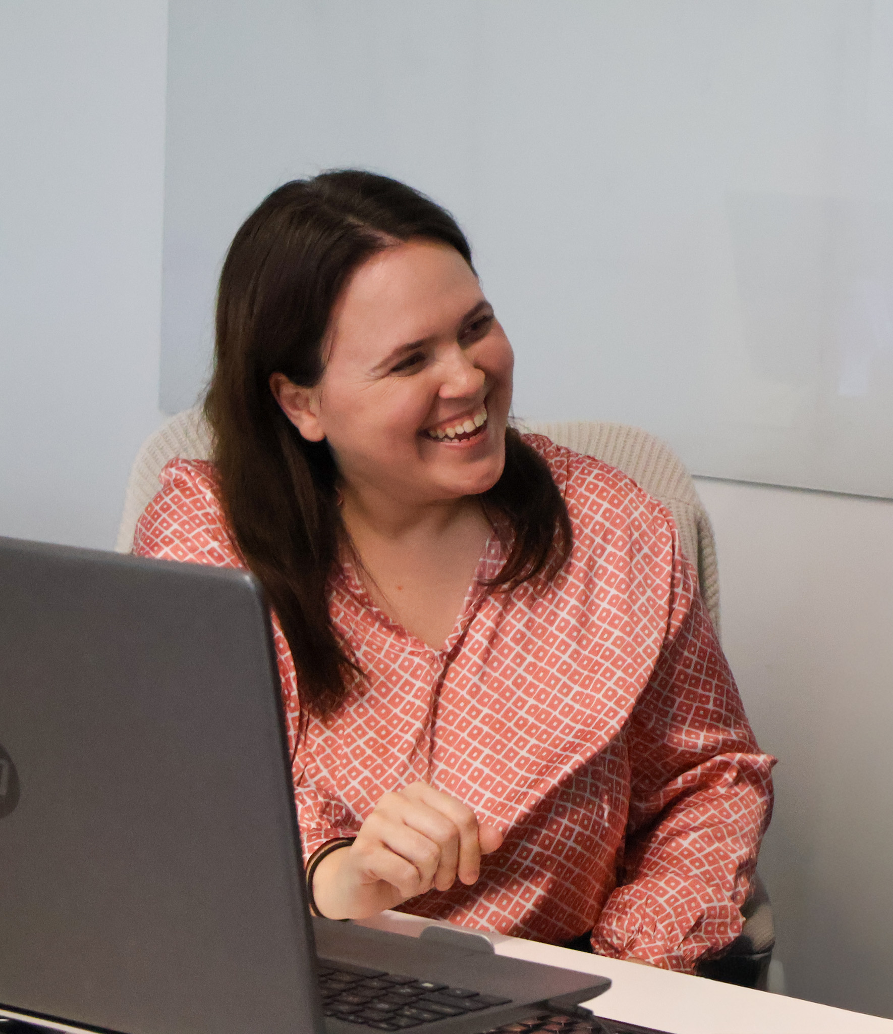 A woman laughing at her desk with a laptop open in front of her.