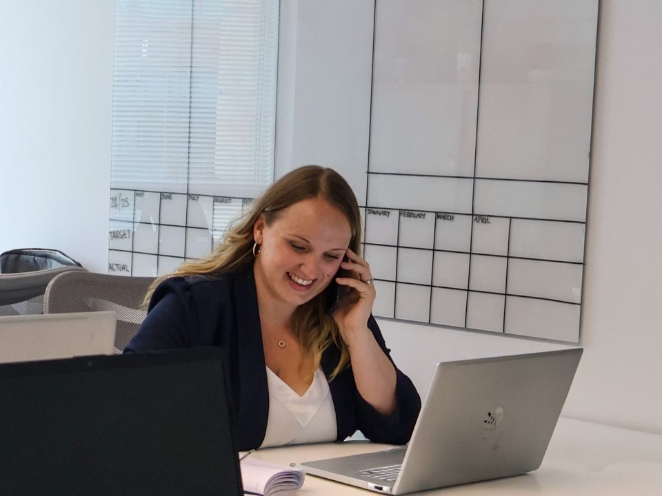 A recruiter smiling and talking on the phone at a desk with a laptop, in a bright office.