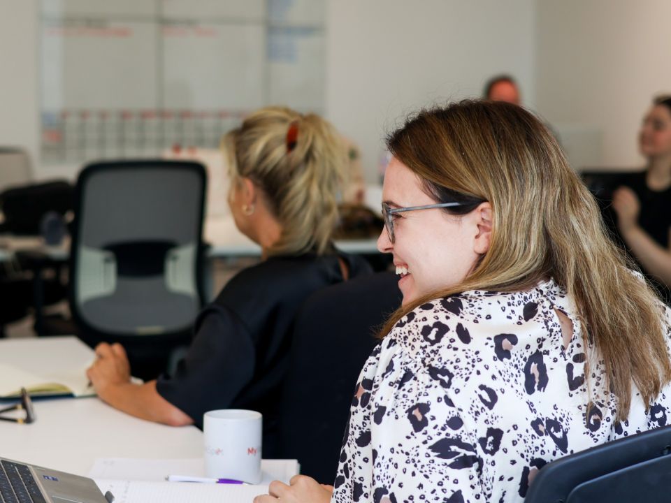 Colleagues smiling and engaged during a team meeting in an open-plan office, with laptops and notebooks on the desk.