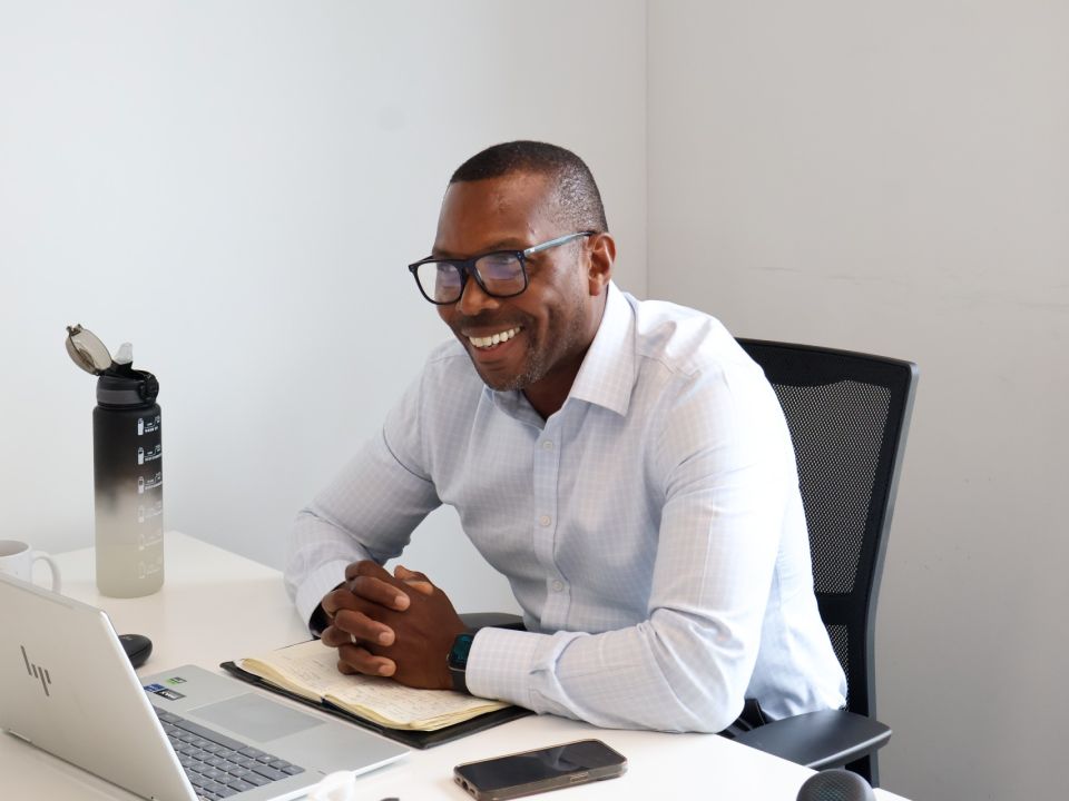 Man smiling at a desk with a laptop, open notebook and phone, mid-conversation during a business meeting.